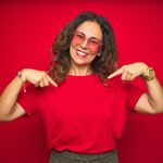 Middle age senior woman wearing cute heart shaped glasses over red isolated background looking confident with smile on face, pointing oneself with fingers proud and happy.