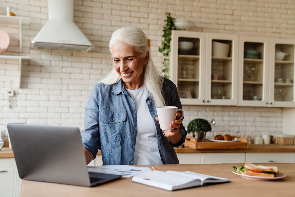 Senior woman smiling while working on her laptop in the kitchen holding cup of tea.